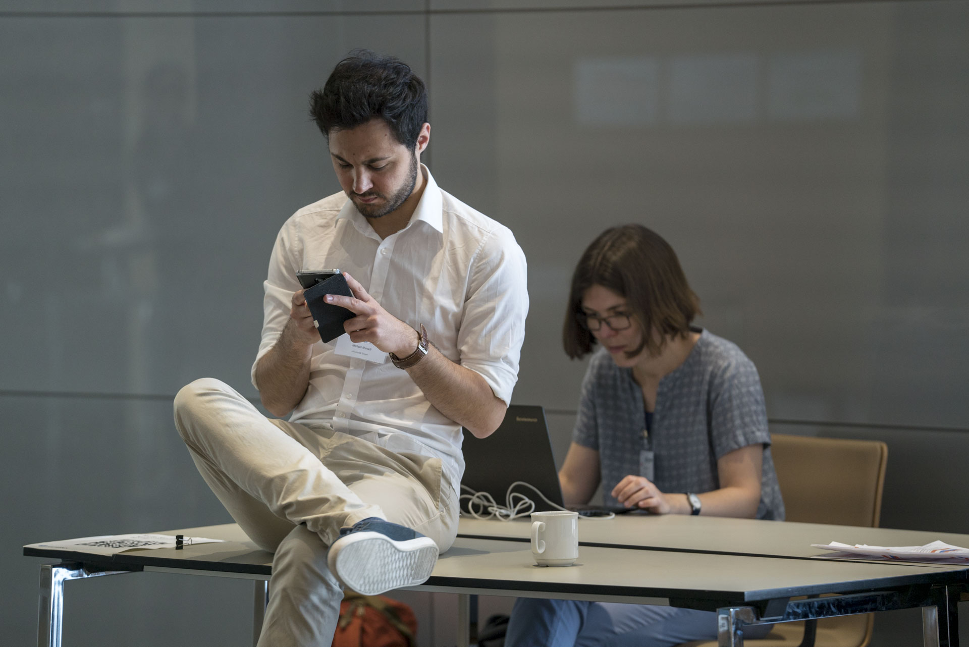 Ein Mann sitzt auf dem Schreibtisch mit Smartphone, eine Frau sitzt am Tisch mit Laptop